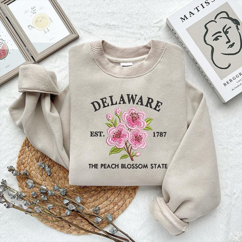 May include: Beige sweatshirt featuring the word "DELAWARE" in black script, embroidered pink flowers, and the text "EST. 1787 THE PEACH BLOSSOM STATE". The sweatshirt has a crew neck and long sleeves.