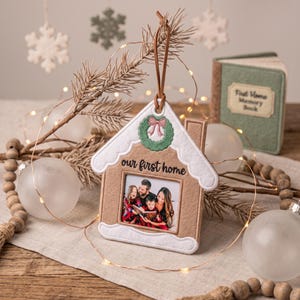 May include: A house-shaped felt Christmas ornament featuring a tan exterior with white scalloped trim, a green embroidered wreath with a pink bow, and the text "our first home" above a square photo frame containing a family portrait.