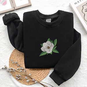 May include: A black long-sleeve sweatshirt featuring a central embroidered design of a white magnolia flower with green leaves. The garment has a ribbed crew neckline and ribbed cuffs at the ends of the sleeves, providing a structured fit.