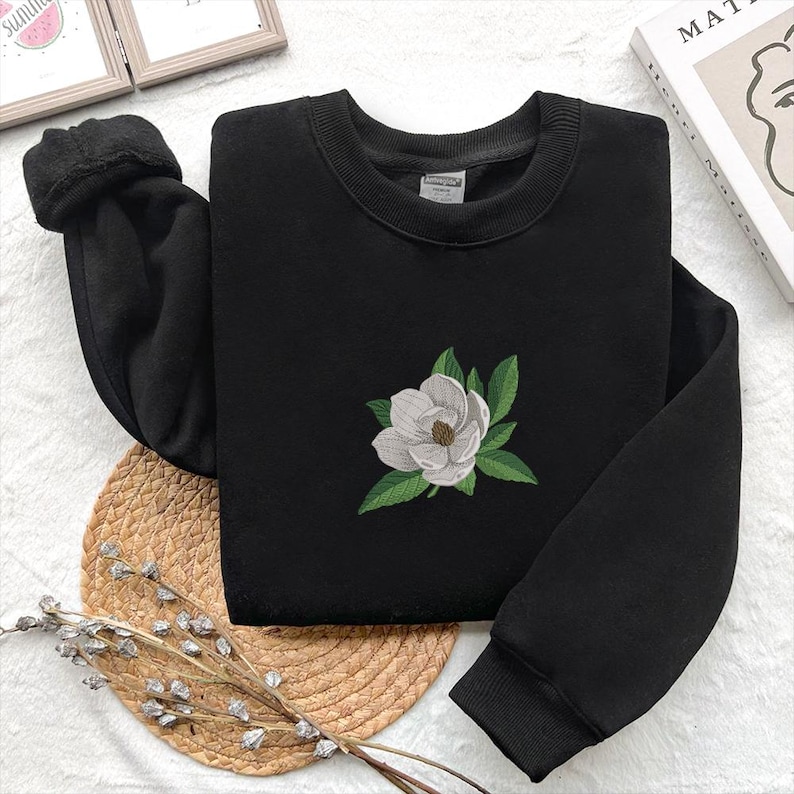 May include: A black long-sleeve sweatshirt featuring a central embroidered design of a white magnolia flower with green leaves. The garment has a ribbed crew neckline and ribbed cuffs at the ends of the sleeves, providing a structured fit.