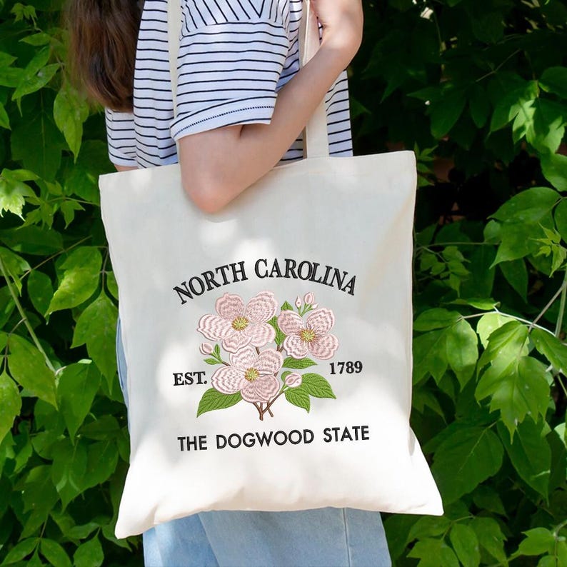 May include: Cream-colored tote bag with embroidered pink dogwood flowers and the text "North Carolina" above the flowers. Below the flowers are the words "Est. 1789" and "The Dogwood State."