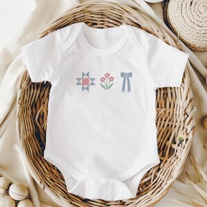 May include: White baby onesie with embroidered designs. The design includes a geometric star, pink flowers, and a blue bow. The onesie is displayed in a woven basket, suggesting a baby clothing item.