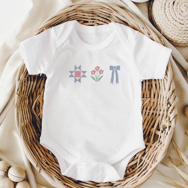 May include: White baby onesie with embroidered designs. The design includes a geometric star, pink flowers, and a blue bow. The onesie is displayed in a woven basket, suggesting a baby clothing item.