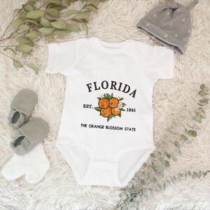 K&ouml;nnte beinhalten: Wei&szlig;er Baby-Body mit dem Wort "FLORIDA" in schwarzer Schrift und einem Orangenbl&uuml;ten-Design. Der Text "EST. 1845 THE ORANGE BLOSSOM STATE" ist ebenfalls auf dem Body aufgedruckt. Graue Babyschuhe, wei&szlig;e Socken und eine graue M&uuml;tze.