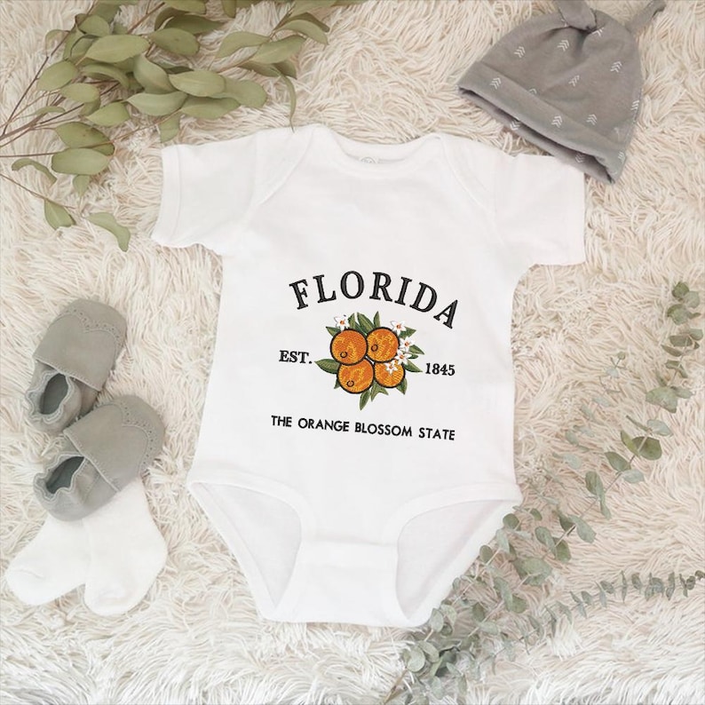 K&ouml;nnte beinhalten: Wei&szlig;er Baby-Body mit dem Wort "FLORIDA" in schwarzer Schrift und einem Orangenbl&uuml;ten-Design. Der Text "EST. 1845 THE ORANGE BLOSSOM STATE" ist ebenfalls auf dem Body aufgedruckt. Graue Babyschuhe, wei&szlig;e Socken und eine graue M&uuml;tze.