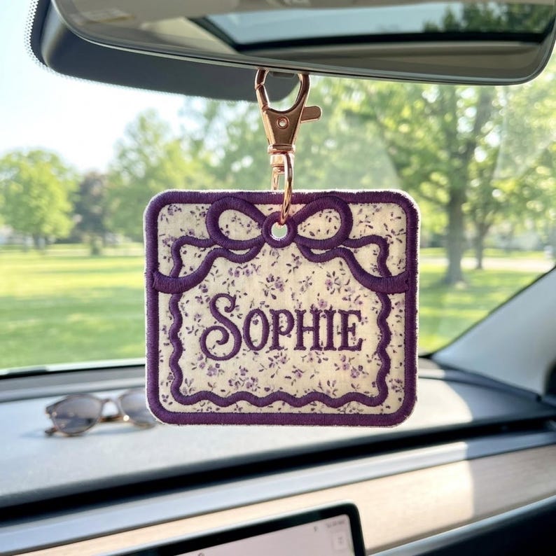May include: A square fabric car charm featuring a white background with a small purple floral pattern. The charm has a dark purple embroidered border shaped like a decorative frame with a bow at the top and the name Sophie embroidered in purple.