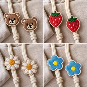 Puede incluir: Una colección de cuatro pares de parches bordados sujetos a cordones trenzados gruesos de color crema. Los diseños incluyen caras de osos marrones, fresas rojas con hojas verdes, margaritas blancas y flores azules con el centro amarillo.