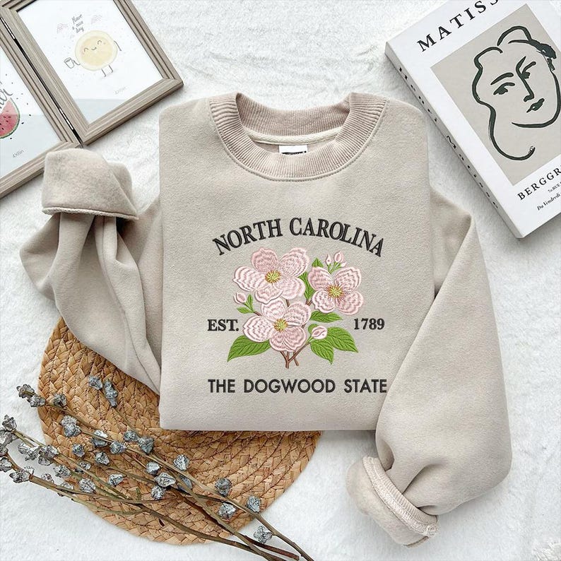 May include: Beige sweatshirt with embroidered floral design and text "NORTH CAROLINA EST. 1789 THE DOGWOOD STATE". The sweatshirt has a crew neck and long sleeves. The design features pink flowers and green leaves.