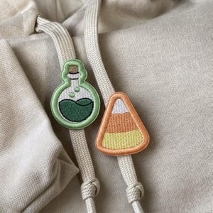 以下が含まれることがあります： ベージュのフード付きスウェットシャツで、ドローストリングに2つの刺繍パッチが付いています。1つはダークグリーンの液体が入ったライトグリーンの魔法の薬瓶、もう1つはオレンジ、イエロー、ホワイトのストライプが入った三角形のキャンディコーンの形をしています。