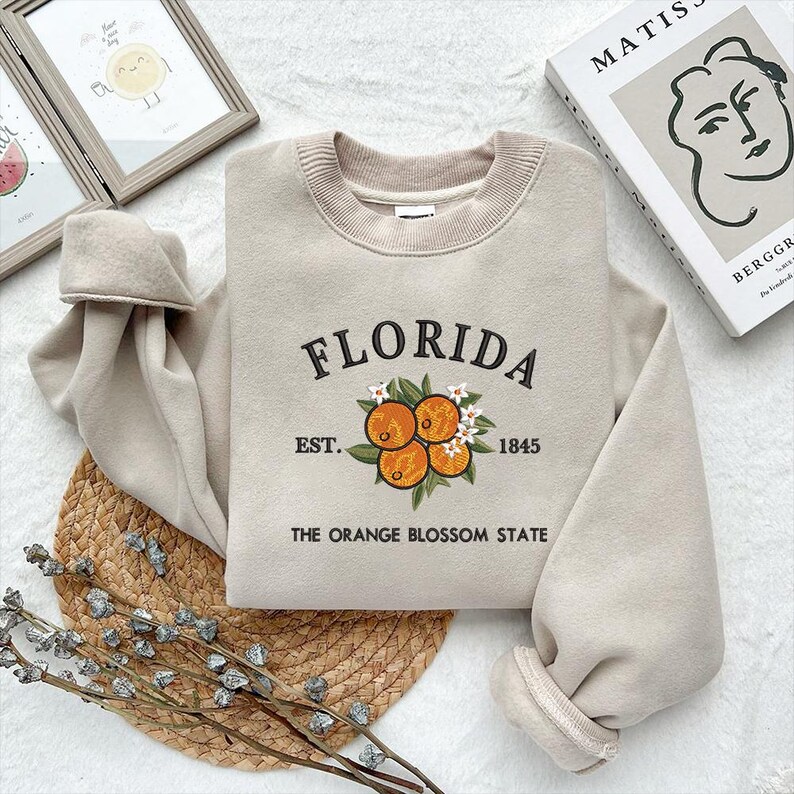 K&ouml;nnte beinhalten: Beiges Sweatshirt mit dem Wort "FLORIDA" in Schwarz gestickt, zusammen mit Orangen und wei&szlig;en Bl&uuml;ten. Der Text "EST. 1845" und "THE ORANGE BLOSSOM STATE" sind ebenfalls gestickt. Das Sweatshirt liegt auf einer gewebten Matte.