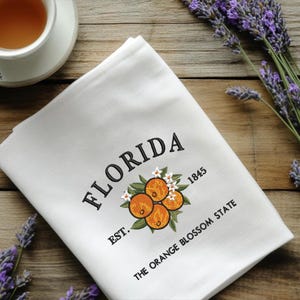 K&ouml;nnte beinhalten: Wei&szlig;es K&uuml;chentuch mit gesticktem Text und Orangenbl&uuml;ten-Design. Das Wort "FLORIDA" ist &uuml;ber einer Ansammlung von Orangen angeordnet, darunter "EST. 1845" und "THE ORANGE BLOSSOM STATE". Eine Tasse Tee und Lavendelzweige sind im Hintergrund.