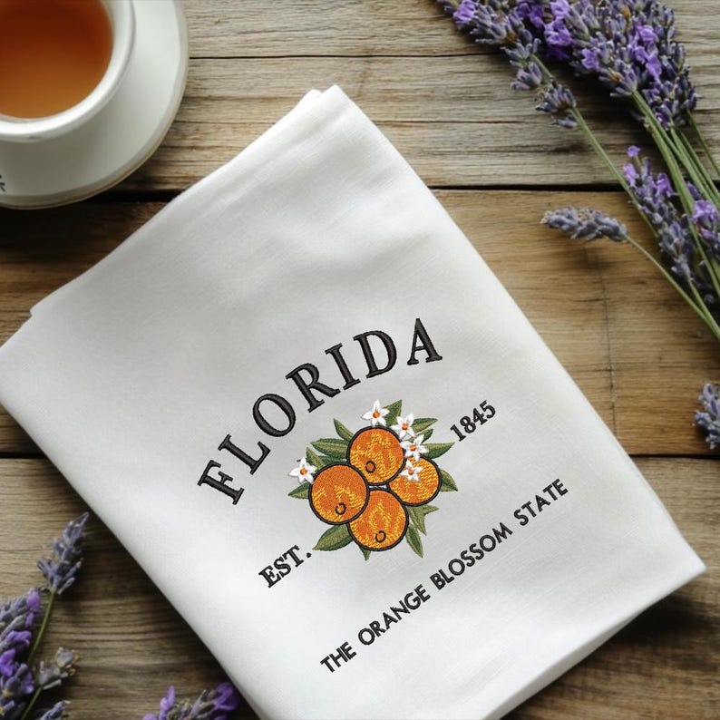 K&ouml;nnte beinhalten: Wei&szlig;es K&uuml;chentuch mit gesticktem Text und Orangenbl&uuml;ten-Design. Das Wort "FLORIDA" ist &uuml;ber einer Ansammlung von Orangen angeordnet, darunter "EST. 1845" und "THE ORANGE BLOSSOM STATE". Eine Tasse Tee und Lavendelzweige sind im Hintergrund.