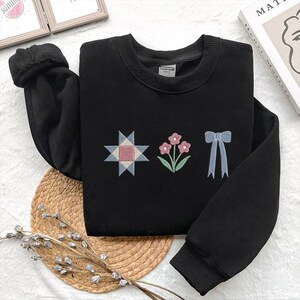 May include: Black sweatshirt with embroidered designs. The designs include a starburst, flowers, and a bow. The starburst is a combination of pink, blue, and white. The flowers are pink with green stems and leaves. The bow is light blue.