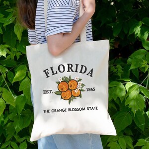 K&ouml;nnte beinhalten: Cremefarbene Tragetasche mit dem Wort "FLORIDA" in schwarzer Schrift. Darunter befindet sich ein gesticktes Design aus Orangen und Blumen. Der Text "EST. 1845" und "THE ORANGE BLOSSOM STATE" sind ebenfalls auf der Tasche aufgedruckt.