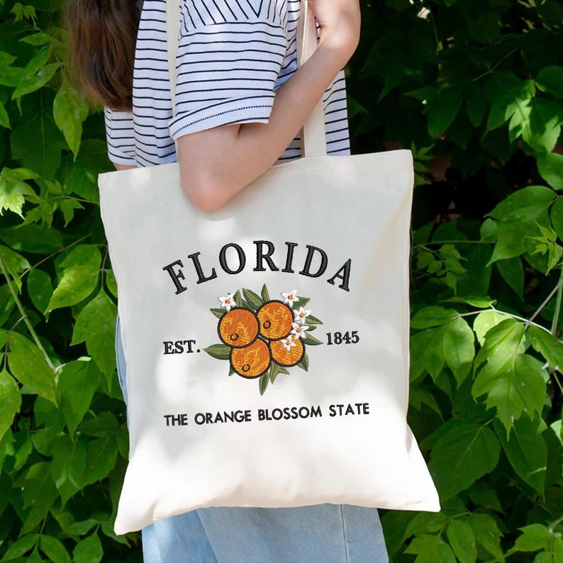 K&ouml;nnte beinhalten: Cremefarbene Tragetasche mit dem Wort "FLORIDA" in schwarzer Schrift. Darunter befindet sich ein gesticktes Design aus Orangen und Blumen. Der Text "EST. 1845" und "THE ORANGE BLOSSOM STATE" sind ebenfalls auf der Tasche aufgedruckt.
