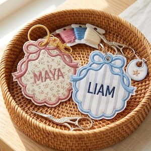 May include: Two scalloped fabric keychains with gold-tone rings and clips. One features a pink floral print with the name MAYA embroidered in pink, and the other has a blue striped pattern with the name LIAM embroidered in blue. Both have bow details.