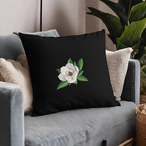 May include: A square black throw pillow featuring a centered embroidered design of a white magnolia flower with a yellow-brown center and detailed green leaves. The embroidery displays visible stitching texture against the solid black fabric.