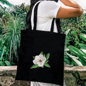 May include: A black tote bag featuring an embroidered design of a white magnolia flower with green leaves centered on the front. The bag has two black shoulder straps and is constructed from a solid black fabric material.