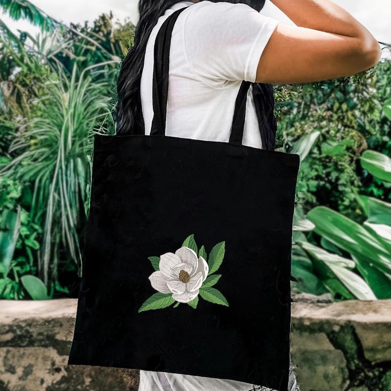 May include: A black tote bag featuring an embroidered design of a white magnolia flower with green leaves centered on the front. The bag has two black shoulder straps and is constructed from a solid black fabric material.