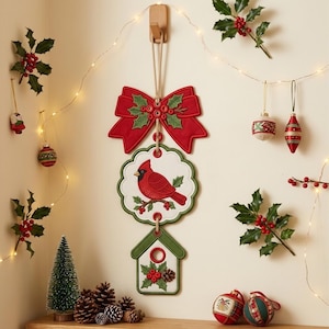May include: A vertical hanging Christmas decoration featuring a red fabric bow with holly leaves at the top, a scalloped white plaque with a red cardinal bird illustration in the center, and a green house-shaped plaque with holly at the bottom.