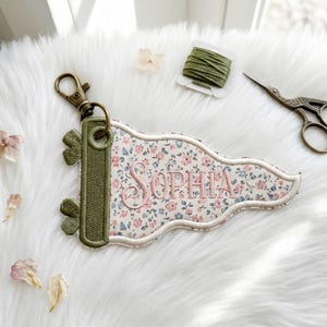 May include: A pennant-shaped keychain featuring a floral pattern in shades of pink and blue on a cream background. The name Sophia is embroidered in pink thread. It has an olive green border, a metal grommet, and a bronze-colored lobster clasp.