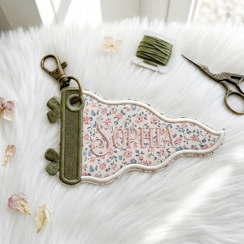 May include: A pennant-shaped keychain featuring a floral pattern in shades of pink and blue on a cream background. The name Sophia is embroidered in pink thread. It has an olive green border, a metal grommet, and a bronze-colored lobster clasp.