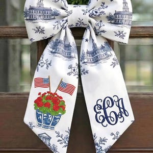May include: A large white bow with a blue and white toile pattern featuring buildings, flowers, and stars. The bow's tails display American flags, red flowers, and a monogram. This decorative bow is perfect for patriotic decor.