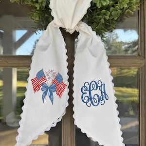 Pode incluir: Faixa de porta branca com bordas onduladas. Um lado apresenta um desenho bordado de bandeiras americanas e fogos de artifício com um laço azul. O outro lado tem um monograma azul. Detalhe decorativo para uma porta ou coroa.