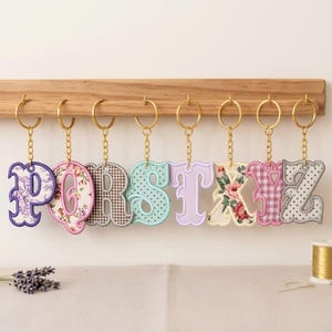 May include: A set of eight embroidered letter keychains featuring the letters P, Q, R, S, T, X, Y, and Z. Each letter is crafted with distinct fabric patterns including floral, gingham, and polka dot designs, attached to gold-toned metal rings.