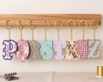 May include: A set of eight embroidered letter keychains featuring the letters P, Q, R, S, T, X, Y, and Z. Each letter is crafted with distinct fabric patterns including floral, gingham, and polka dot designs, attached to gold-toned metal rings.