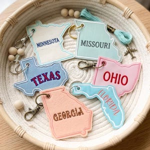 Puede incluir: Una colección de seis llaveros con forma de estados de EE. UU. con texto bordado para Minnesota, Missouri, Texas, Ohio, Georgia y Florida. Cada llavero incluye un cierre de mosquetón de metal y está dispuesto dentro de una cesta de cuerda.