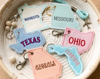 May include: A collection of six state-shaped keychains featuring embroidered text for Minnesota, Missouri, Texas, Ohio, Georgia, and Florida. Each keychain includes a metal lobster clasp and is arranged inside a round, woven rope basket.