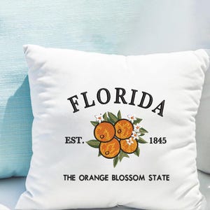 K&ouml;nnte beinhalten: Wei&szlig;es quadratisches Kissen mit gesticktem Design. Das Design zeigt das Wort "FLORIDA" &uuml;ber einer Ansammlung von Orangen, Bl&auml;ttern und Bl&uuml;ten. Darunter stehen die Worte "EST. 1845" und "THE ORANGE BLOSSOM STATE".