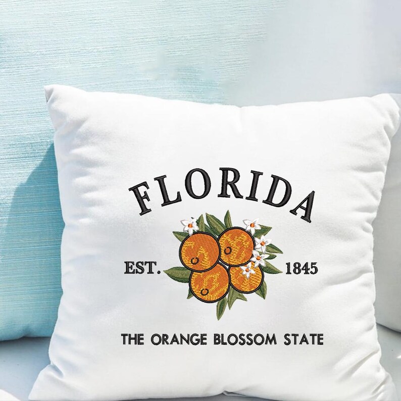 K&ouml;nnte beinhalten: Wei&szlig;es quadratisches Kissen mit gesticktem Design. Das Design zeigt das Wort "FLORIDA" &uuml;ber einer Ansammlung von Orangen, Bl&auml;ttern und Bl&uuml;ten. Darunter stehen die Worte "EST. 1845" und "THE ORANGE BLOSSOM STATE".