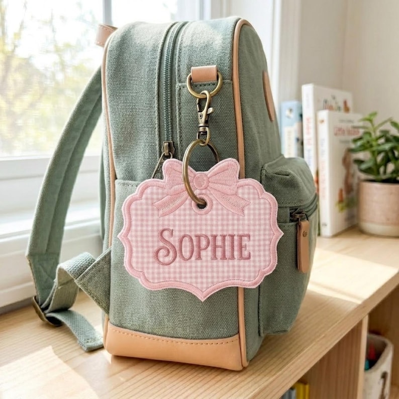May include: A sage green canvas backpack with tan trim features a hanging luggage tag. The tag is a scalloped, light pink and white gingham fabric shape with an embroidered bow at the top and the name SOPHIE stitched in dark pink thread.