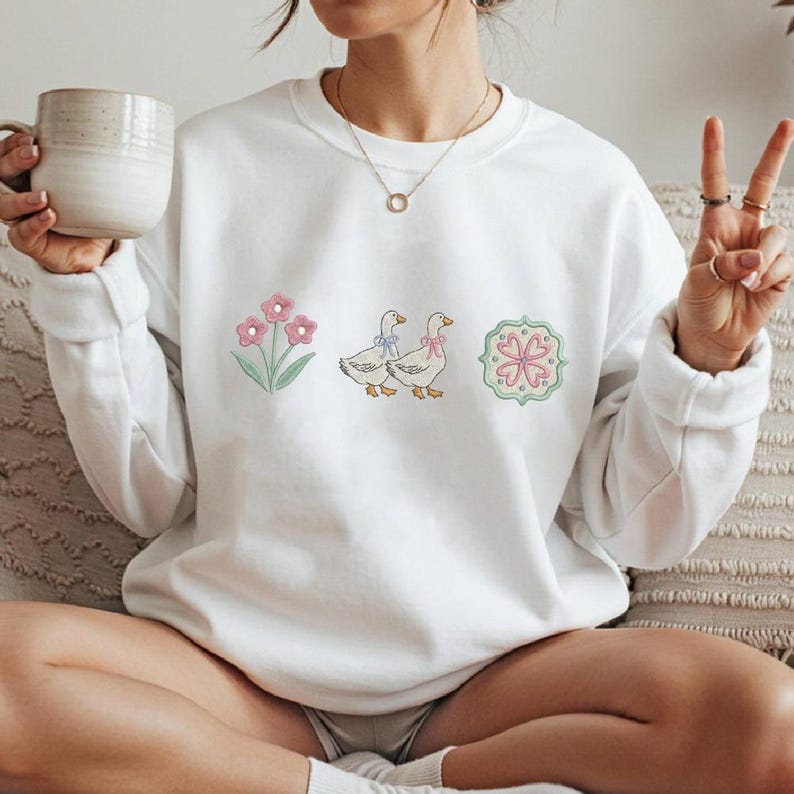 May include: White sweatshirt with embroidered floral, goose, and heart designs. The floral design features pink flowers with green stems. Two geese with blue and pink bows are next to a heart-shaped design with pink and green accents. A casual, comfortable style.