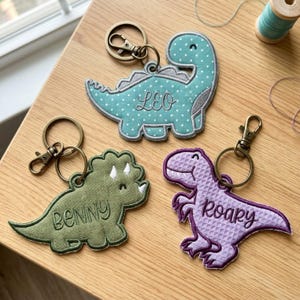 Puede incluir: Tres llaveros de dinosaurio bordados con cierres de mosquetón de metal. Un dinosaurio de cuello largo color verde azulado con puntos blancos y el nombre LEO, un tricerátops verde con el nombre BENNY y un dinosaurio morado texturizado con el nombre ROARY.