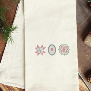 May include: Off-white kitchen towel with three embroidered designs: a pink and blue starburst, a blue oval with a pink heart, and a pink and green floral pattern. The towel is folded and has a simple, clean design.