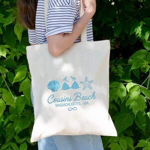 Puede incluir: Bolso tote de color crema con un dise&ntilde;o azul de concha, top de bikini y estrella de mar. El texto "Cousins Beach Massachusetts, USA" est&aacute; impreso debajo, con un s&iacute;mbolo de infinito. El bolso es llevado.