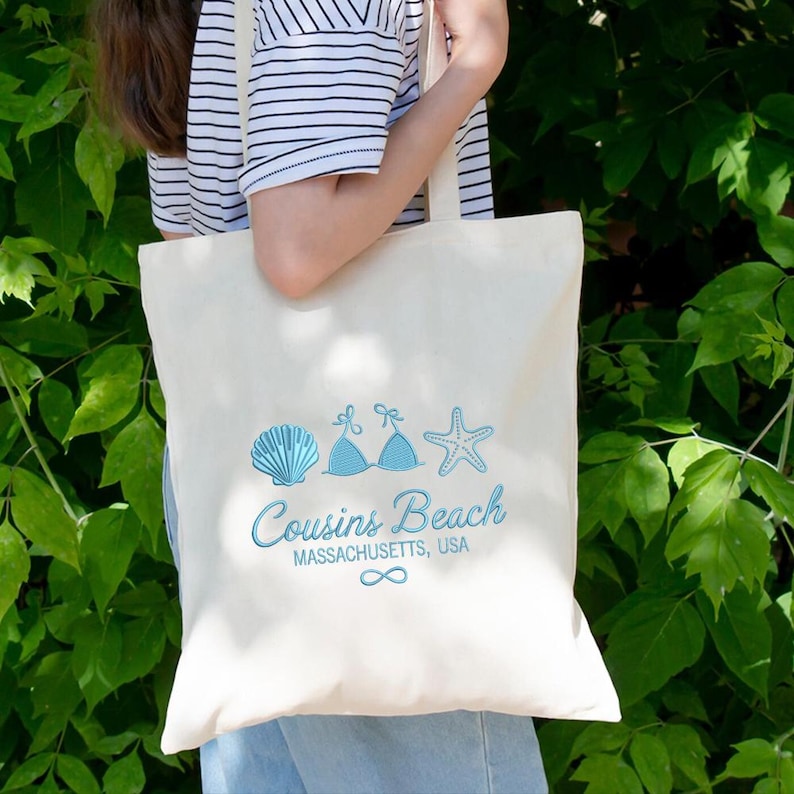 Puede incluir: Bolso tote de color crema con un dise&ntilde;o azul de concha, top de bikini y estrella de mar. El texto "Cousins Beach Massachusetts, USA" est&aacute; impreso debajo, con un s&iacute;mbolo de infinito. El bolso es llevado.