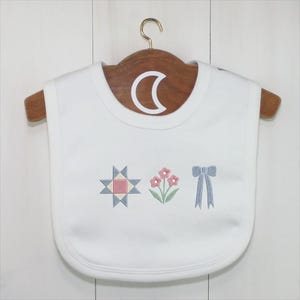 May include: White baby bib with embroidered designs. The bib features a star, flowers, and a bow in pastel colors. The bib is hanging on a wooden hanger.