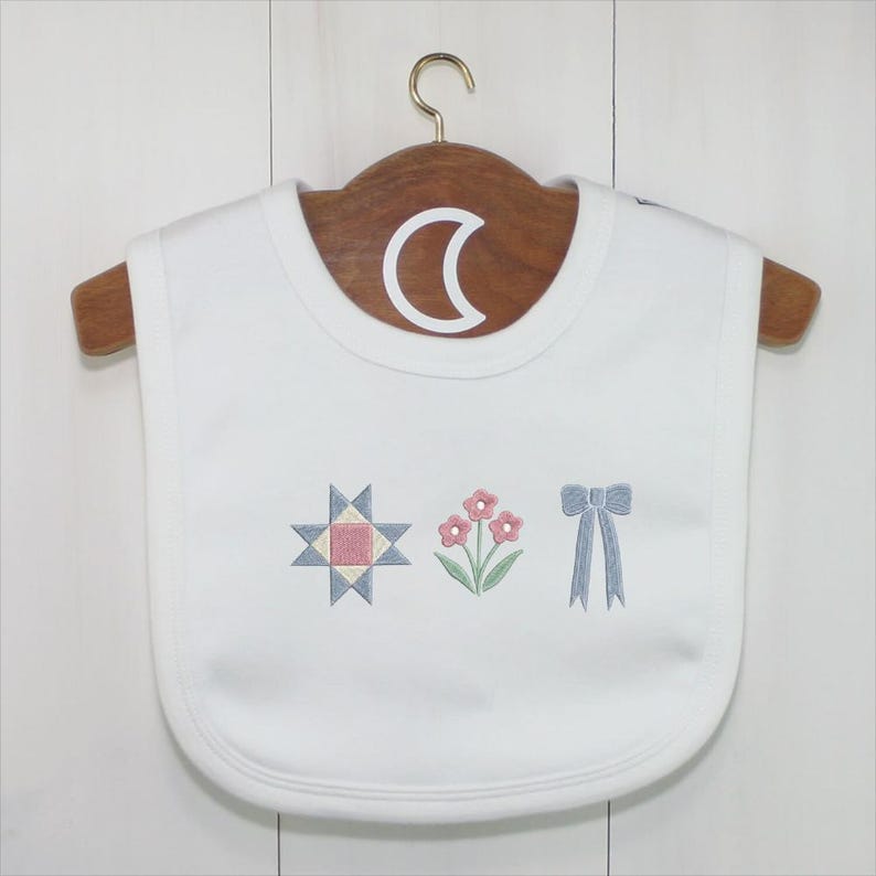 May include: White baby bib with embroidered designs. The bib features a star, flowers, and a bow in pastel colors. The bib is hanging on a wooden hanger.