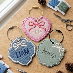 May include: Three embroidered luggage tags with metal rings. One pink heart-shaped tag with the name ARIA and a bow, one blue scalloped tag with the name LIAM and a bow, and one sage green scalloped tag with the name MAYA and a bow.