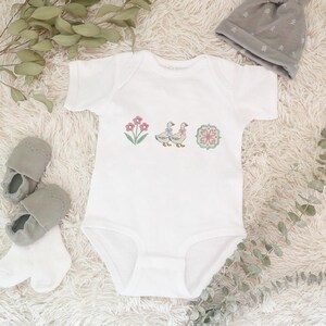 May include: White baby onesie with embroidered floral and goose designs. The onesie features a pink flower, two geese with blue bows, and a decorative floral emblem. Gray baby shoes, socks, and a gray beanie are also in the image.