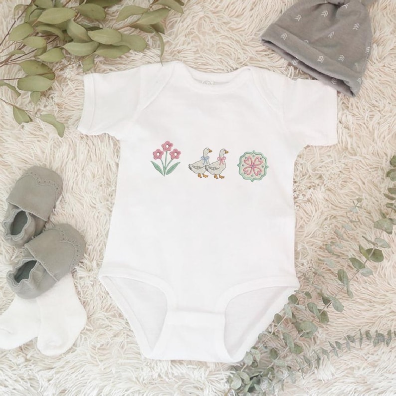 May include: White baby onesie with embroidered floral and goose designs. The onesie features a pink flower, two geese with blue bows, and a decorative floral emblem. Gray baby shoes, socks, and a gray beanie are also in the image.