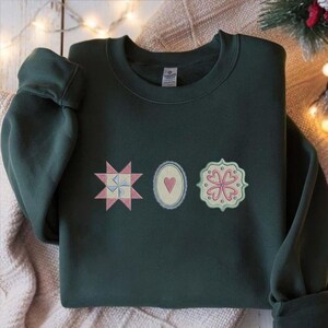 May include: Dark green sweatshirt with three embroidered patches. The patches include a pink and blue starburst, a heart in an oval, and a floral design. The sweatshirt is folded on a beige blanket.