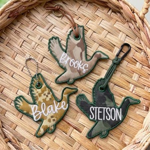 May include: Three duck-shaped keychains with embroidered names. One features a tan camouflage pattern with the name Brooks, one a pixelated tan pattern with the name Blake, and one a dark green camouflage pattern with the name STETSON.