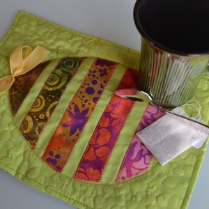 Peut inclure: Un dessous de tasse matelassé vert avec un motif d'œuf coloré et un ruban jaune. Une tasse verte et un sachet de thé sont sur le dessous de tasse.