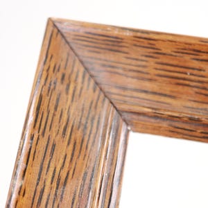 May include: Close-up of a corner of a wooden picture frame. The frame is made of brown wood with visible grain and a natural finish. The corner is joined at a 90-degree angle, showcasing the craftsmanship of the frame.