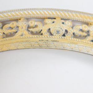 May include: A curved, decorative frame segment featuring a pale yellow finish with distressed edges revealing a grey base. The piece displays intricate, raised scrollwork, floral motifs, and a rope-like border pattern along the outer edge.